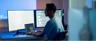 Masada oturan ve kod görüntüleyen iki büyük monitörün bağlı olduğu bir dizüstü bilgisayarda çalışan bir kişi.