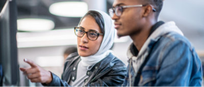 Birlikte oturan, dikkatle odaklanmış iki kişi. Birisi bir şeye işaret ediyor ve her ikisi de gündelik bir ortamda gözlük takıyor.