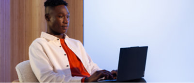 Hafif aydınlatılmış bir odada masada oturan ve dizüstü bilgisayar kullanan beyaz ceketli ve kırmızı gömlekli bir kişi.