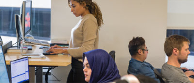 A person standing in front of a table with a person using a computer