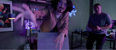 A man with long hair, glasses, and a beard reaching out to the side, wearing a purple shirt and black pants.