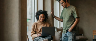 A man in a green shirt stands next to a woman wearing headphones and using a laptop.