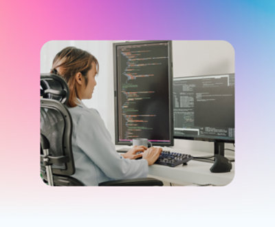 A person in a grey shirt, seated on an office chair, types code on a keyboard in front of two large computer