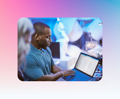 A person in a blue shirt is holding a laptop and showing its screen to another person in a blurred background.