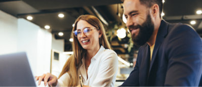 Two professionals, a woman and a man, smiling and looking at a laptop screen together in a modern office setting.