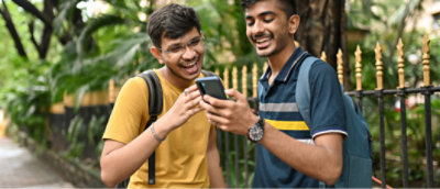 Two young men laughing and looking at a smartphone together, standing outdoors with greenery in the background.