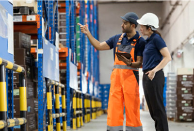 A man in orange overalls holding a tablet next to a woman in a blue shirt in a warehouse.