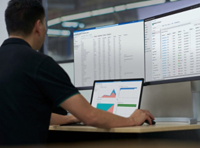 Seorang pria mengenakan kaus hitam bekerja menggunakan laptop dengan beberapa monitor komputer yang menampilkan teks.