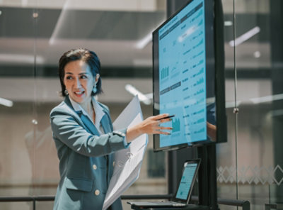 Seorang wanita dengan setelan biru menunjuk ke arah layar.