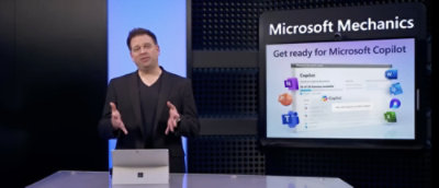 Seorang pria dengan setelan hitam berdiri di depan layar yang menampilkan Microsoft Mechanics: Bersiap untuk Microsoft Copilot.