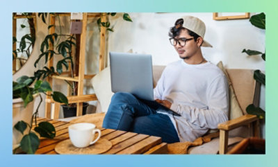 A man wearing a white shirt and glasses is seated on a couch, working on a laptop.