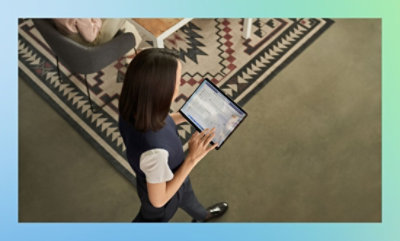 A person is holding and using a tablet while walking on a patterned rug next to a table.