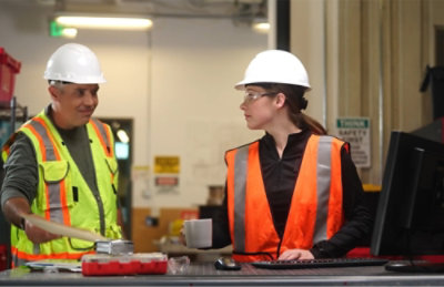 ヘルメットと安全ベストを着用した 2 人の建設作業員が、ツールとコンピューターが置かれた作業台の前で会話をしています。