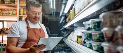 一名穿著橙色圍裙的男子，在一排食品架前手持平板電腦。