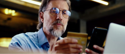 A man with glasses and a beard looks at a gold card in one hand and holds a smartphone in the other.