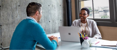 Two individuals in a meeting with one person facing away from the camera and the other working on a laptop at a table.