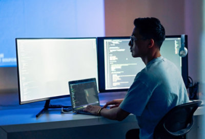 Person working on a laptop with dual monitors showing code in a modern workspace.