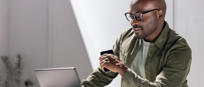 A man with glasses smiles at a tablet while holding a smartphone and wearing a green jacket.
