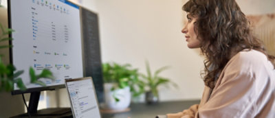 A woman sitting at a desk looking at a laptop and a large monitor screen.