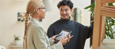 Two people talk in a modern office. One holds a tablet with graphs; the other leans on a wooden shelf, smiling.