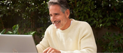 A man in a white sweater smiling at the camera with a laptop.