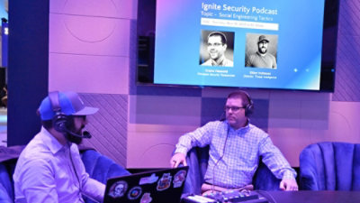 Two men wearing headphones and sitting in a chair in front of a screen displaying Ignite Security Podcast.