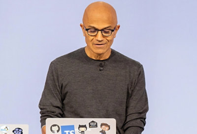 Person speaking at a laptop with stickers, wearing glasses and a grey sweater against a plain background.
