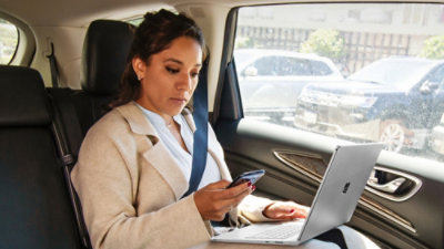 Una persona sentada en el asiento trasero de un automóvil usando un portátil y mirando un smartphone