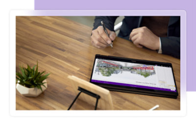 Una persona escribe con un lápiz en una tableta que muestra un diseño arquitectónico, junto a una pequeña planta sobre un escritorio de madera.