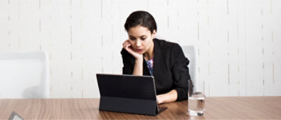 Una mujer con americana negra se sienta en una mesa y utiliza una tableta. Un vaso de agua está colocado sobre la superficie de madera junto a ella.