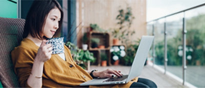 Una persona sentada en un balcón con plantas, sosteniendo una taza y utilizando un portátil.