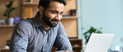 Un hombre con barba y camisa gris sonríe mientras mira un portátil en una oficina.