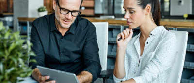 A man and a woman are engaged in a discussion while looking at a tablet in a modern office setting.