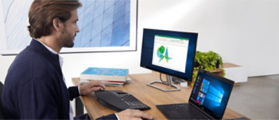 A person works at a desk using a laptop and a desktop monitor displaying charts. A stack of books and a plant are in the background.