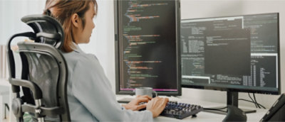 A woman seated at a desk, working on two monitors with a keyboard in front of her, focused on her tasks.