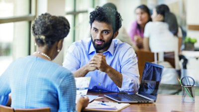 A man and woman sitting at a table, talking, with a laptop in front of them.