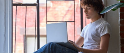 Persoon die een wit t-shirt draagt, op een vensterbank zit en een laptop gebruikt.