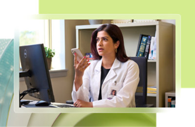 En doktor i en hvit frakk sitter ved et skrivebord og snakker i telefonen. En datamaskin, bøker og en lampe er på skrivebordet.