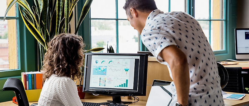 Two people working together in an office, reviewing charts and graphs displayed on a computer monitor.