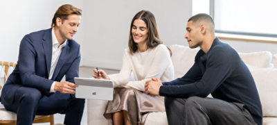 Three colleagues sit together on a sofa in a modern office setting, discussing work around a tablet computer.