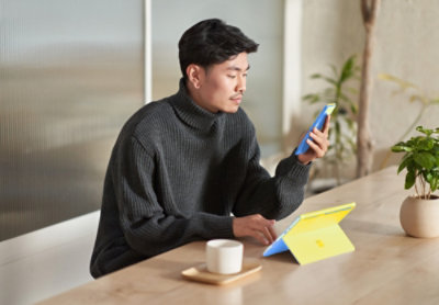 Person sitting at a table using a tablet with a keyboard case while looking at a smartphone, with a cup and plant nearby.