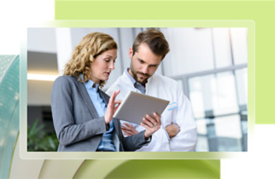 A woman in a business suit and a man in a lab coat are discussing information on a tablet in a bright office setting.