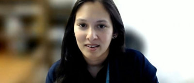 A person with long dark hair wearing a dark top is looking towards the camera with a neutral expression in a blurred indoor setting.