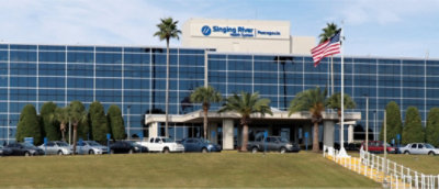 Front view of Singing River Health System hospital building with glass facade, palm trees, parked cars, and a U.S. flag on a pole.
