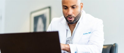 A bearded man in a white coat looking at a laptop.