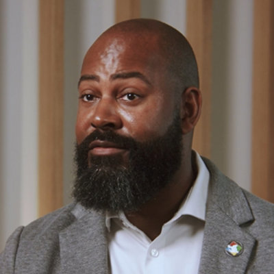 Portrait of a bearded person in a gray blazer with a lapel pin.