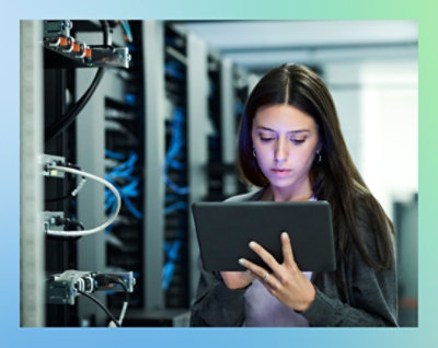A woman stands in a server room, holding a tablet while examining the equipment around her.