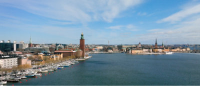 A city with buildings and a tower next to a body of water with boats in a harbor under a blue sky with clouds.