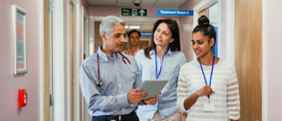 A group of people standing in a hospital hallway, including a woman holding a tablet and a man with a stethoscope