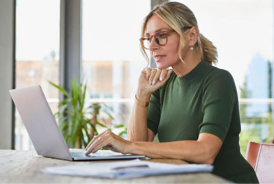 A woman in a green sweater works on a laptop at a wooden table, surrounded by documents, with windows in the background.